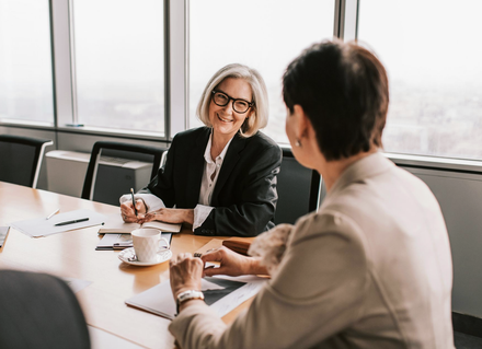 Two coworkers in a bright conference room discussing over coffee and papers