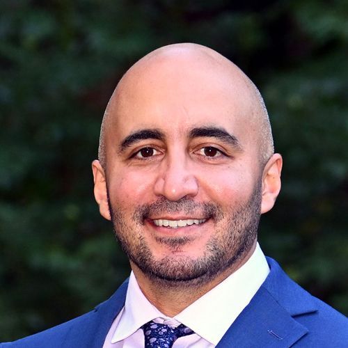 Bald man wearing a blue suit, white shirt, and patterned tie, smiling outdoors.