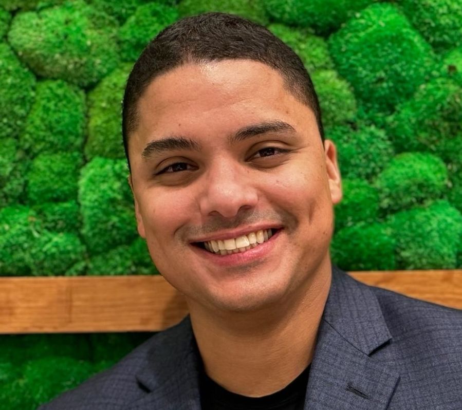 Man smiling, dark hair, wearing a blazer, in front of a green moss wall.
