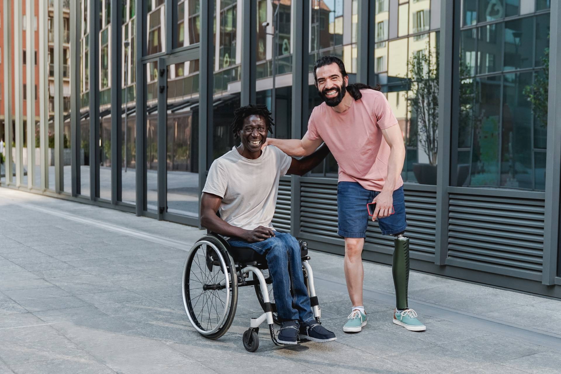 A person using a prosthetic leg and a person in a wheelchair on a city sidewalk.
