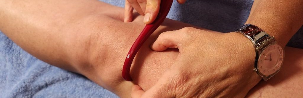 Eine Person massiert sich mit einem roten Werkzeug das Bein. Das Bein liegt auf einem blauen Tuch. Eine Uhr ist zu sehen.