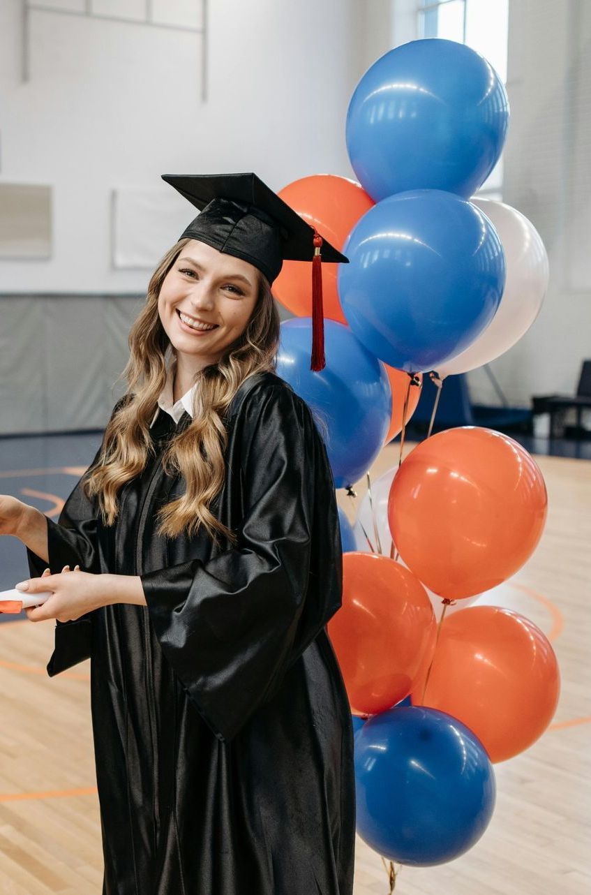 A woman in a graduation cap and gown is standing next to a bunch of balloons.