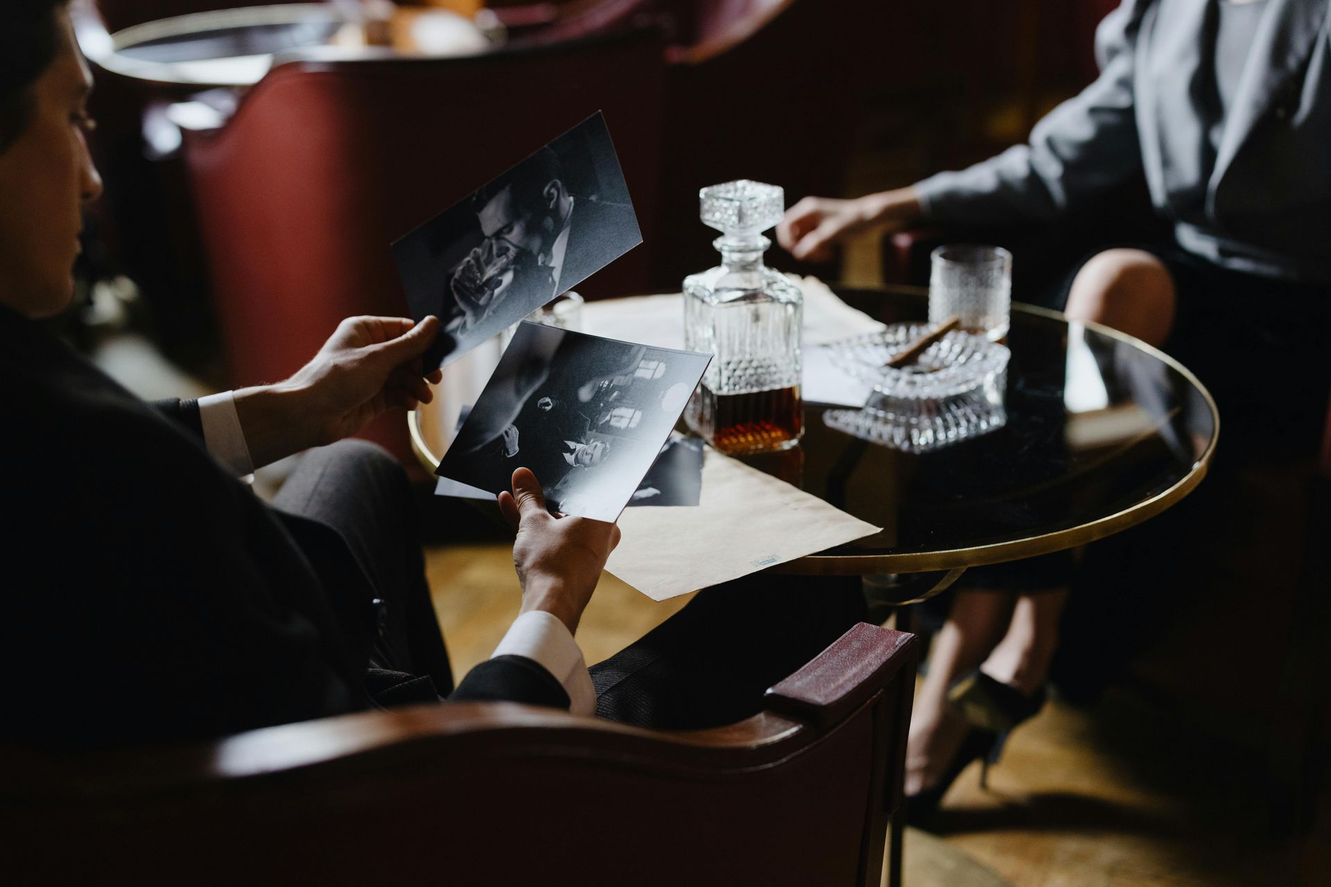 Man in a suit examines photos at a table with a drink, cigar, and person in background.