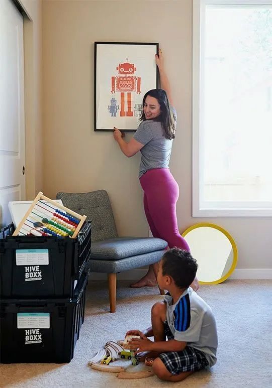 A Woman Is Hanging A Picture On A Wall While A Boy Sits On The Floor — Local & Country Removals in Helensvale, QLD