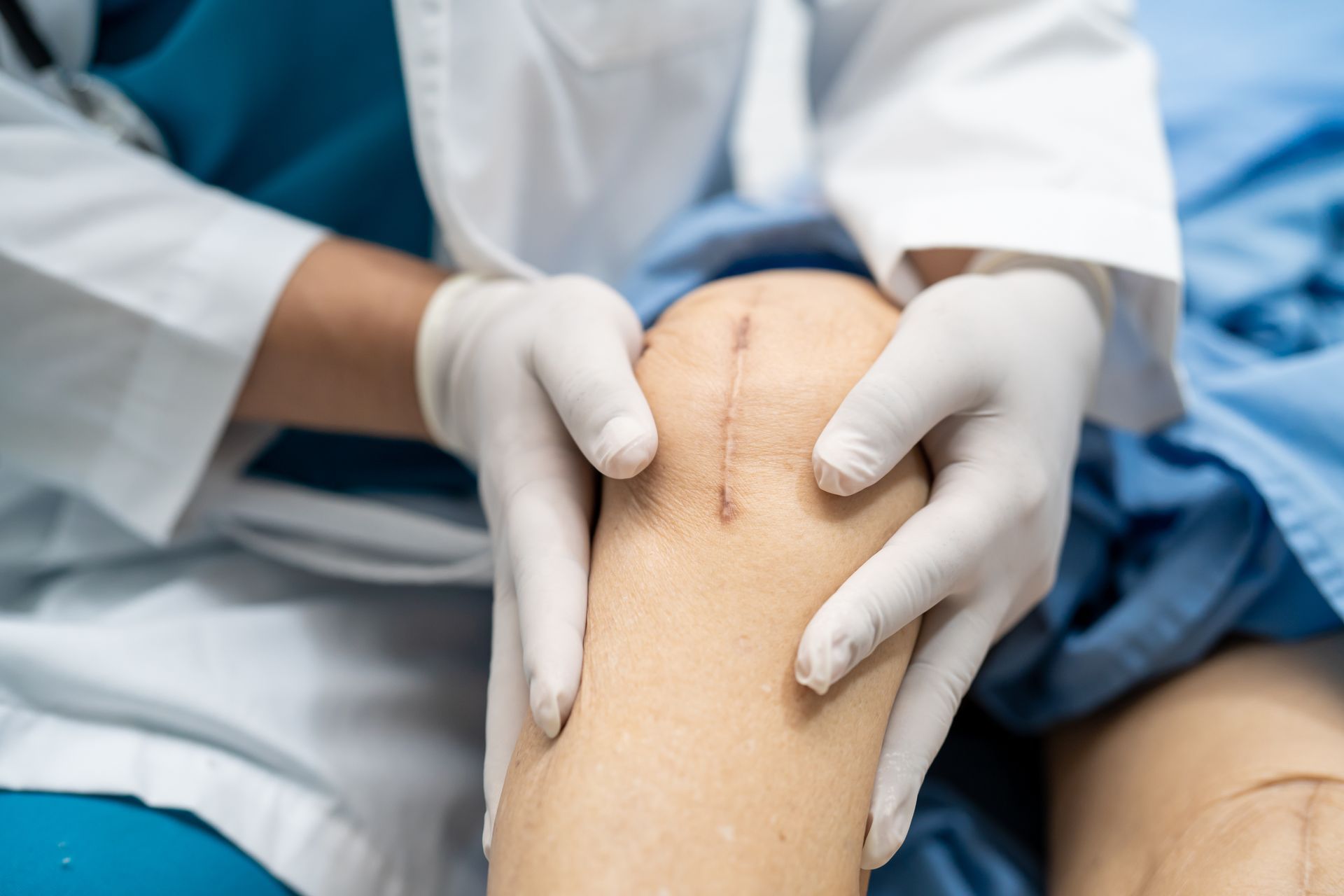 Doctor examines a knee with a surgical scar, wearing gloves, in a hospital setting.