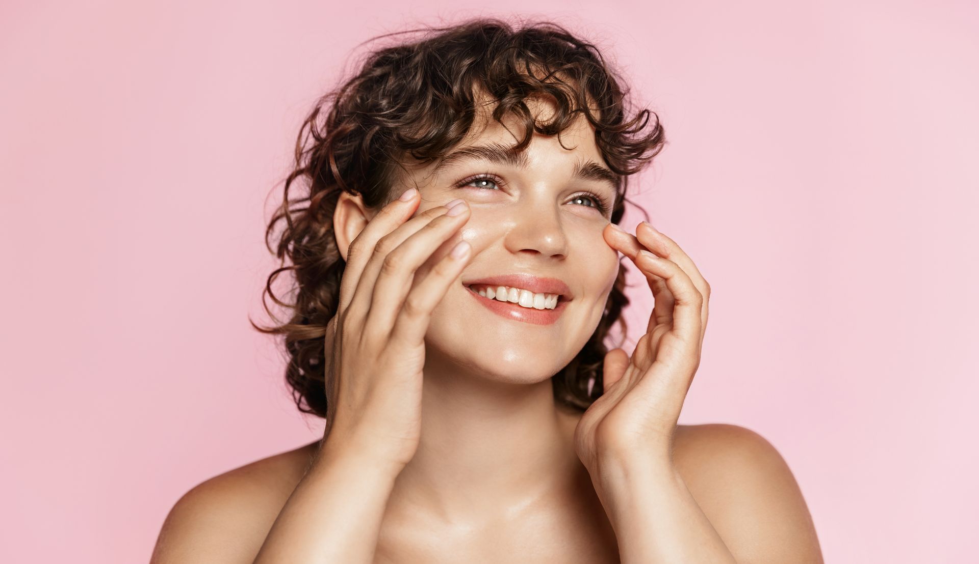 Woman applying skincare product, smiling with pink backdrop.