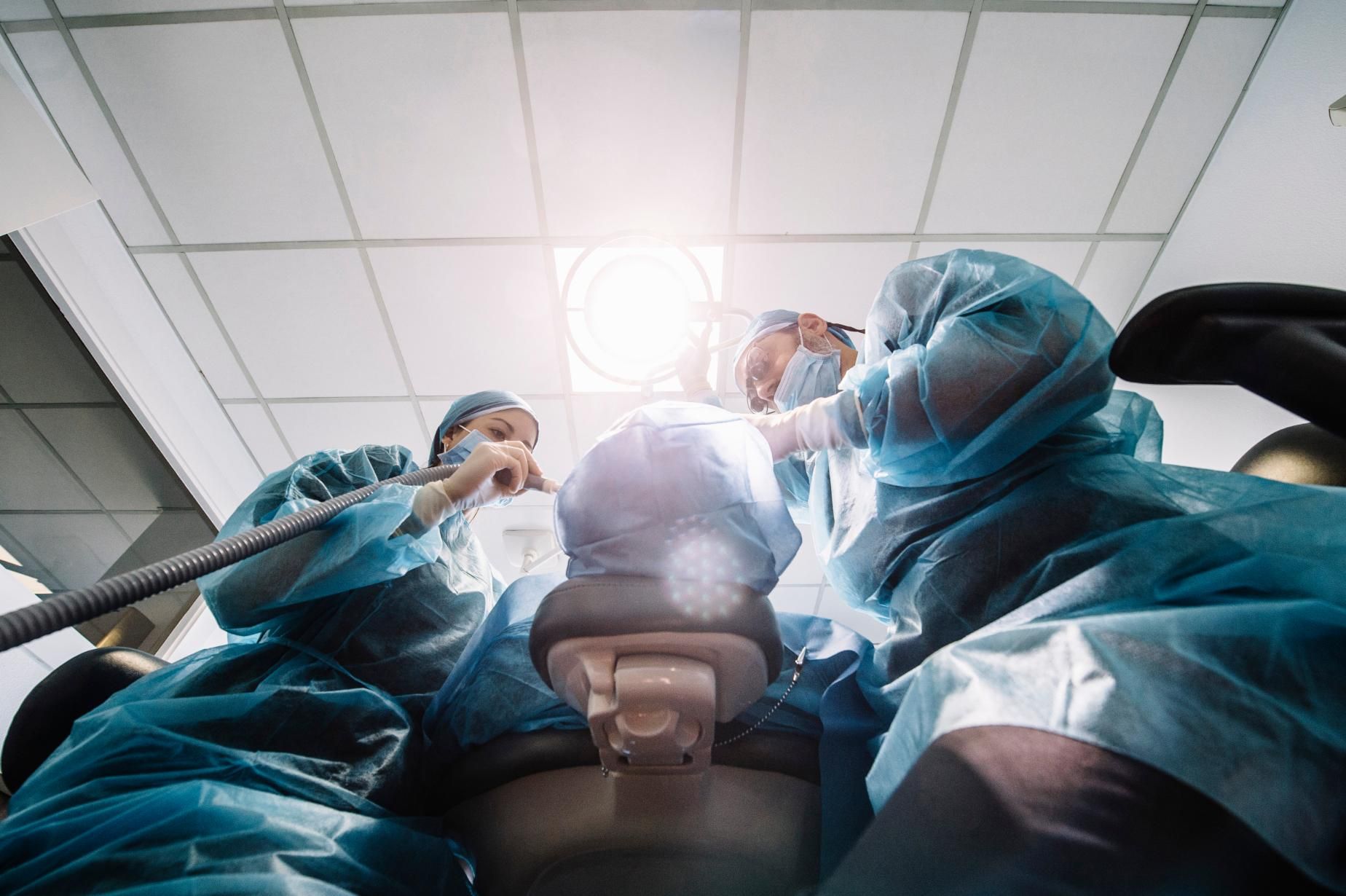 Dentists in blue surgical gowns working around a patient in a dental clinic, viewed from below.