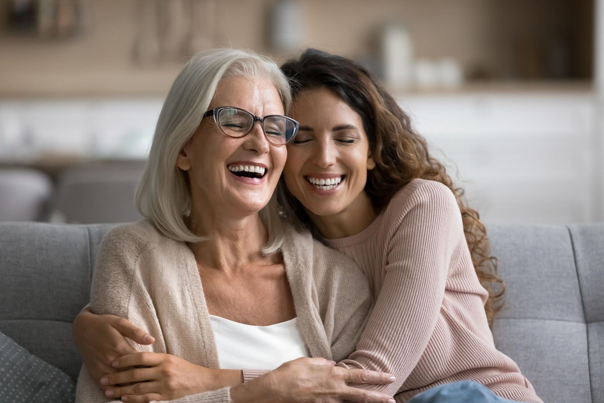 Two women smiling and hugging on a gray couch in a cozy living room