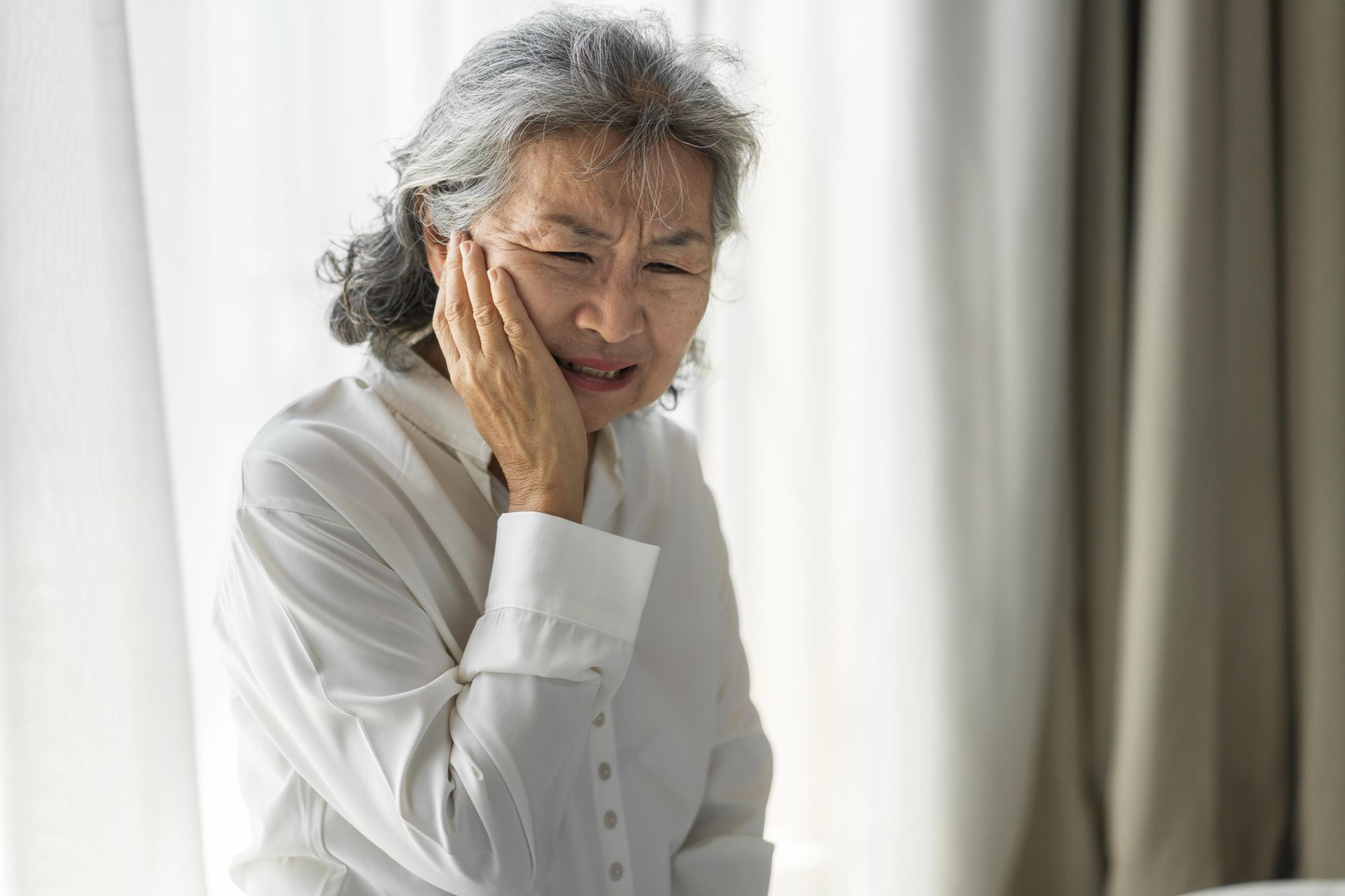 Elderly person in a white shirt holding their cheek, grimacing near a bright window.