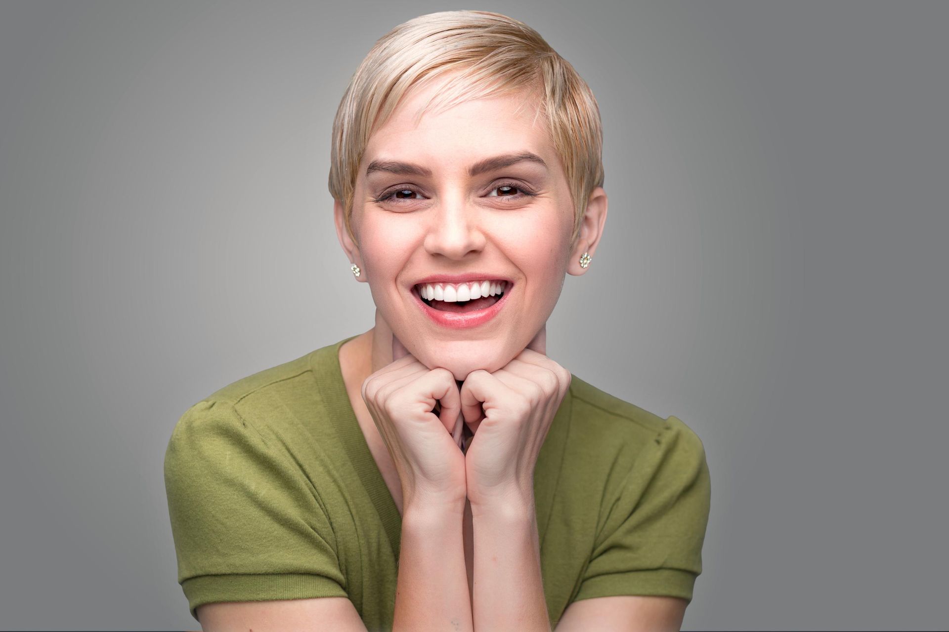 Smiling person with short blonde hair, resting chin on hands, wearing a green shirt against a gray background