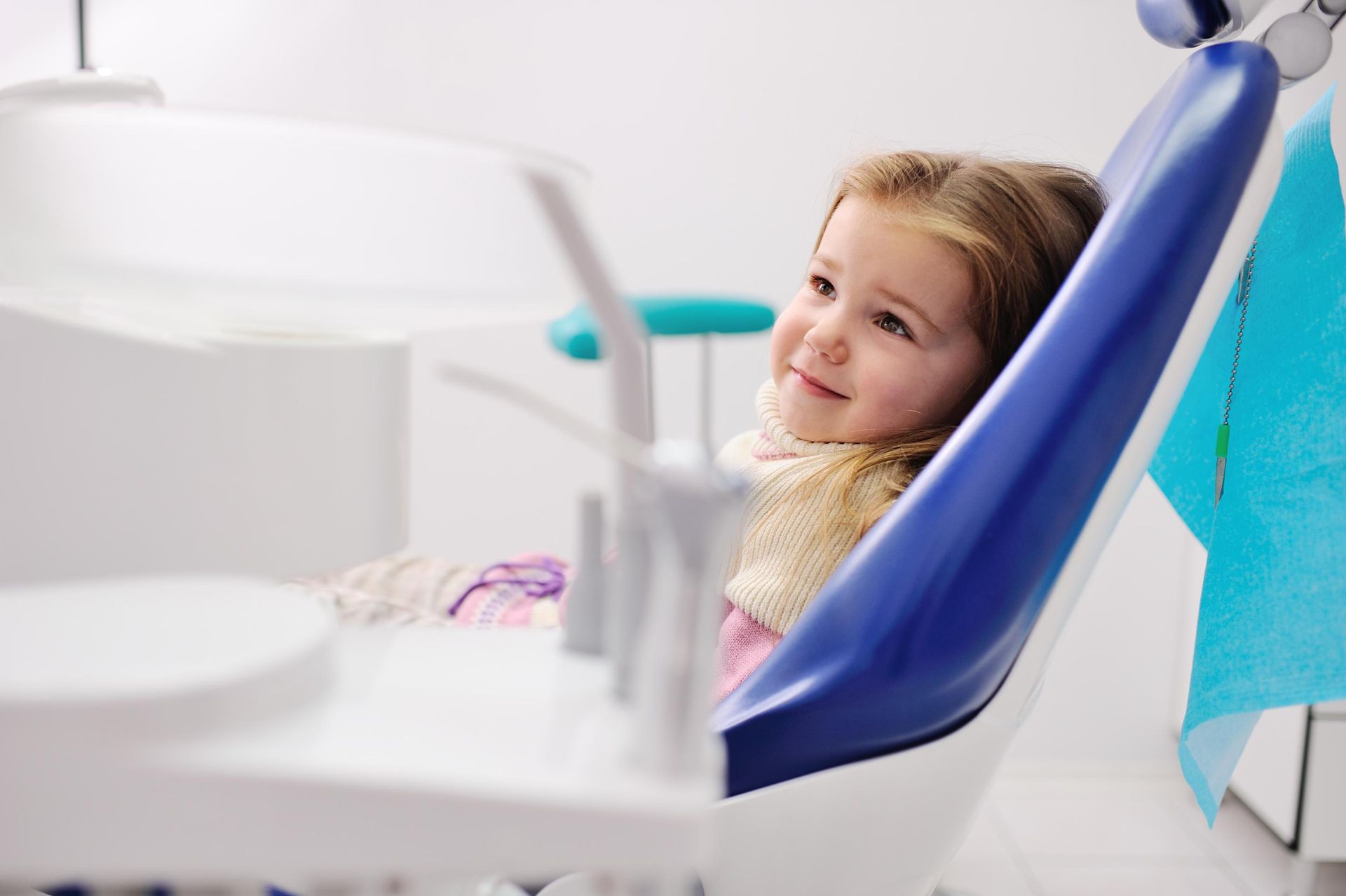 Smiling child reclining in a blue dental chair at a bright clinic, with dental tools in the foreground