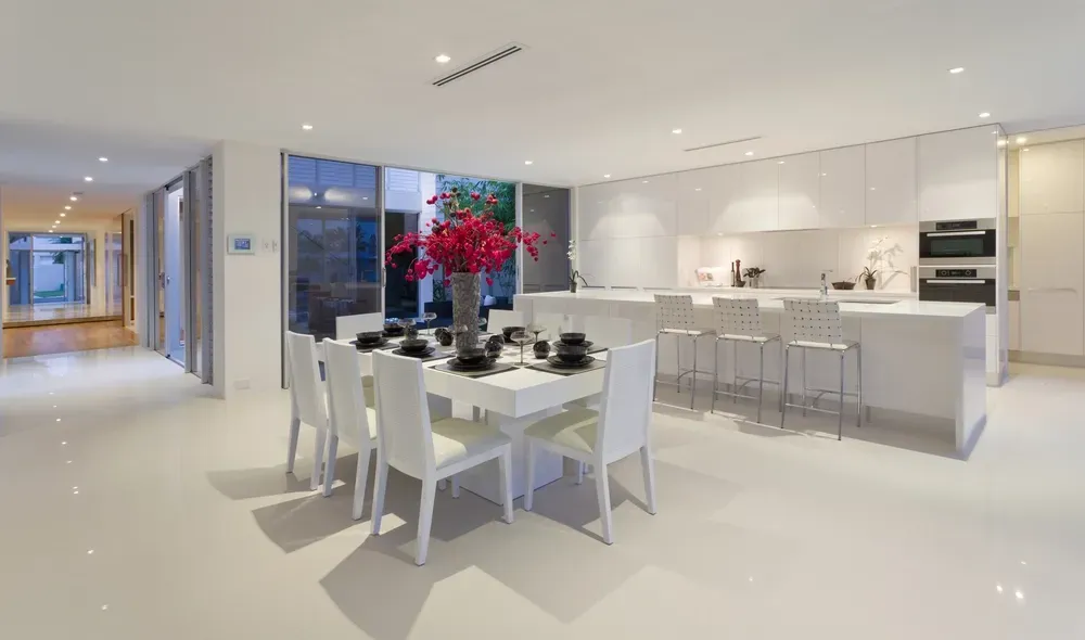 Modern white kitchen and dining area with a table, island, and large windows showing a bright interior.