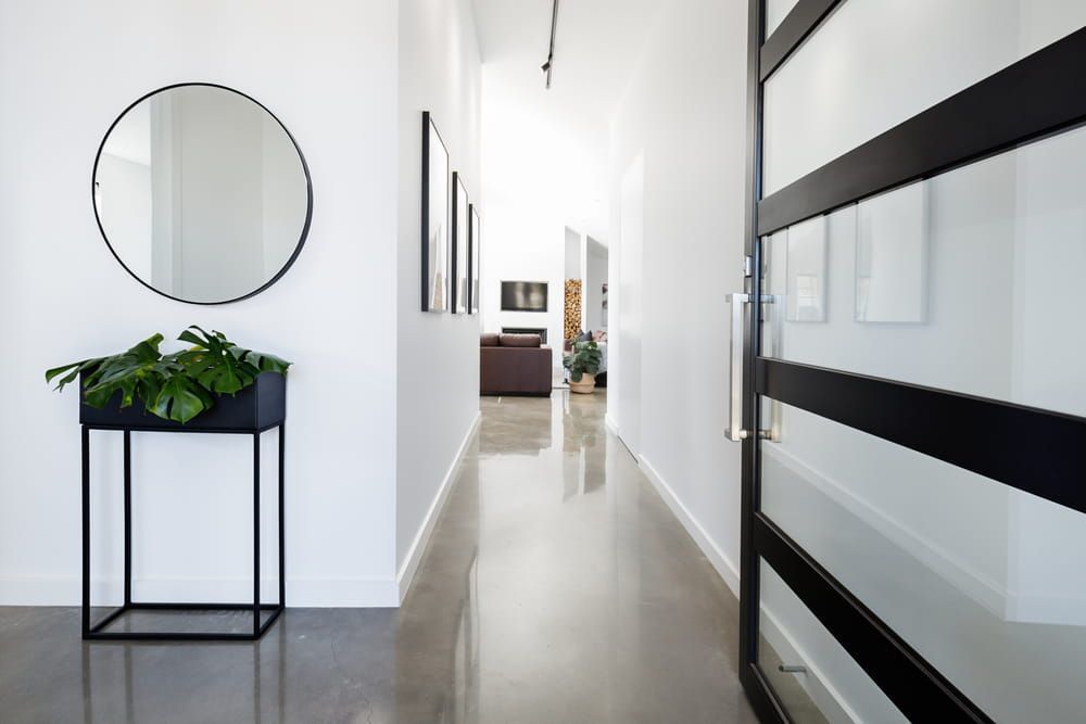 A modern hallway with polished concrete floors, a round mirror over a black console table, and a black-framed glass door.