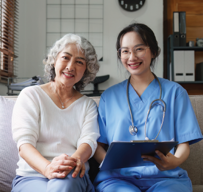 Woman and medical professional smiling, sitting on a couch. The medical professional wears scrubs and holds a clipboard.