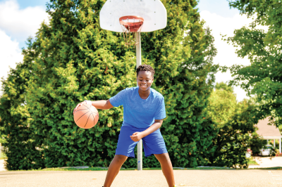 A person dribbles a basketball on a court near a hoop and trees. They wear blue and smile.