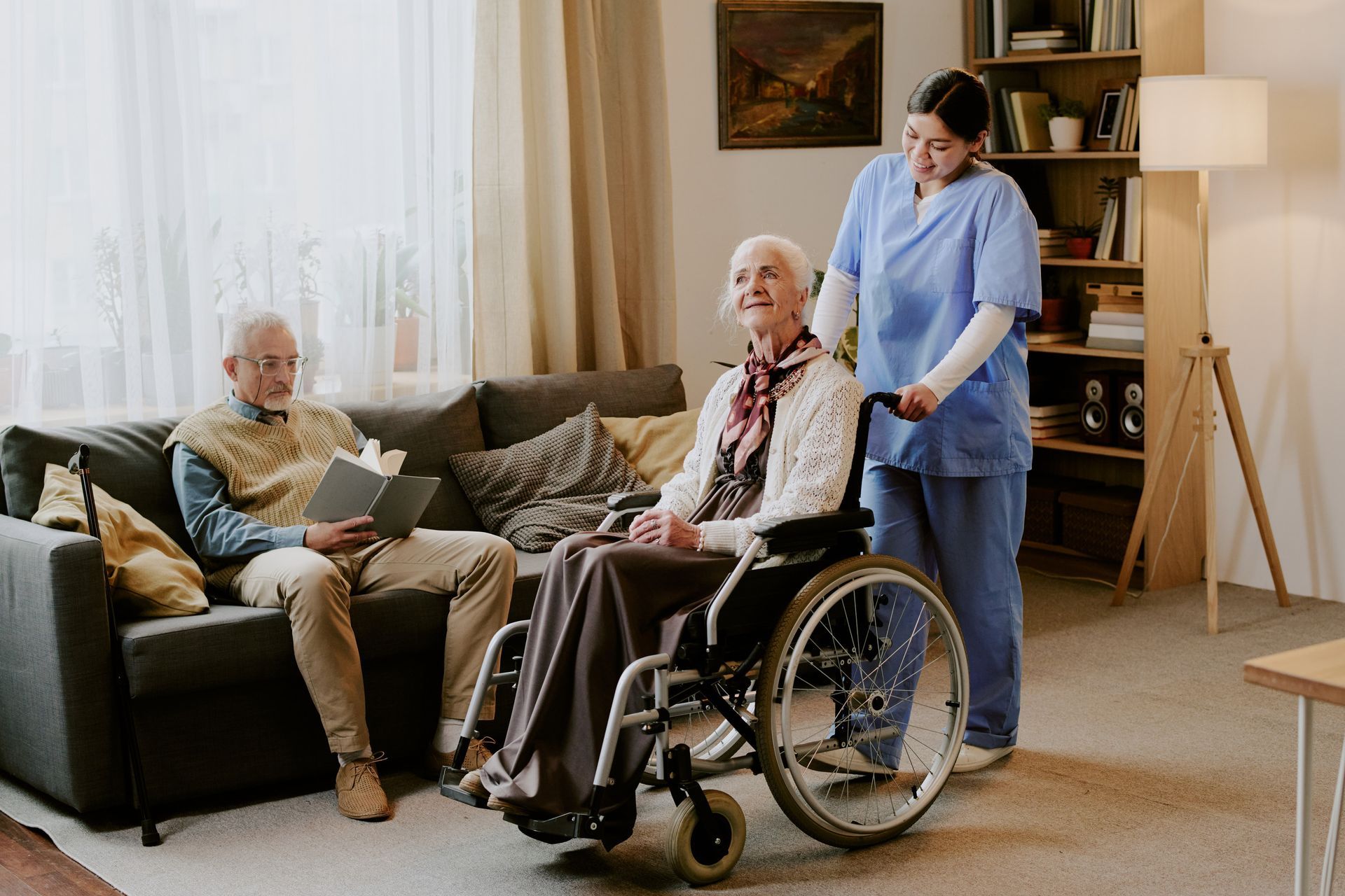 Woman in wheelchair with caregiver, man on sofa. Indoor living room scene.