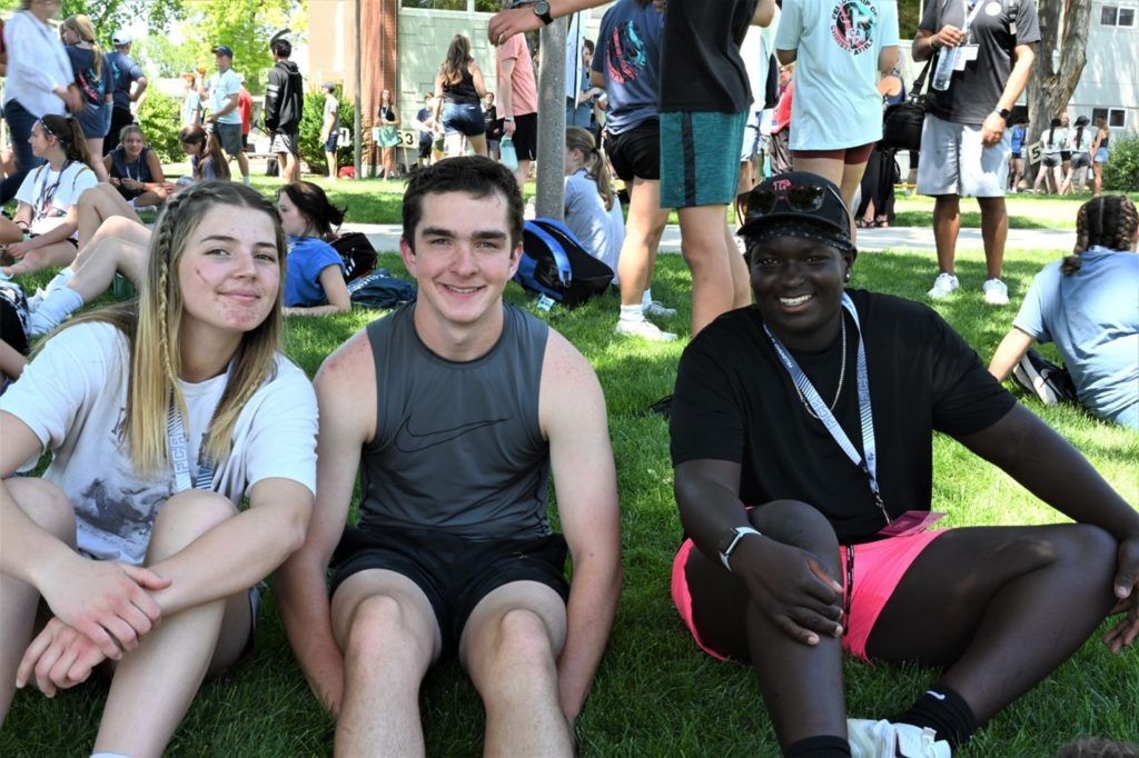 Three people sitting on grass smiling, others in background.