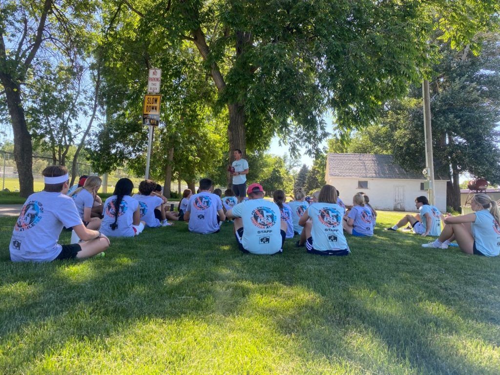 Group of people in light blue shirts and headbands sitting on grass under a tree, listening to a person standing.