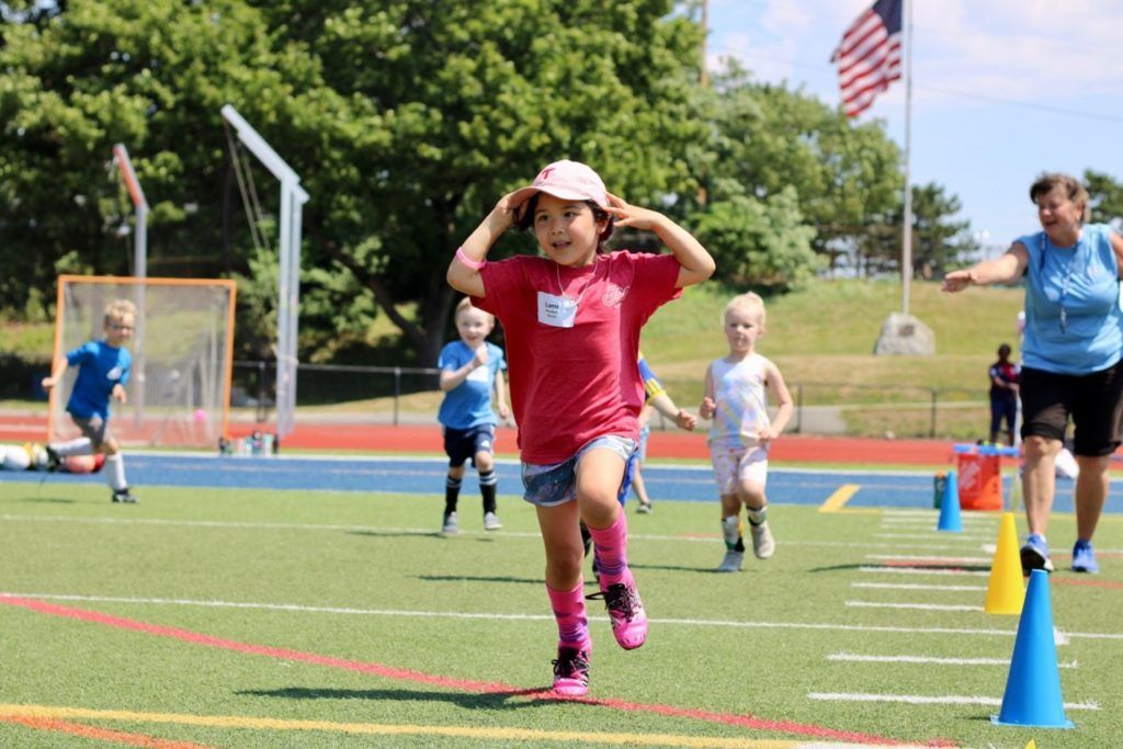 Girl running on a track, arms raised. Others run in the background. Blue sky, American flag.