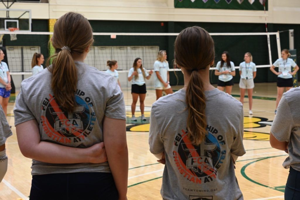 Two people in gray shirts with logos face a volleyball team in a gym.