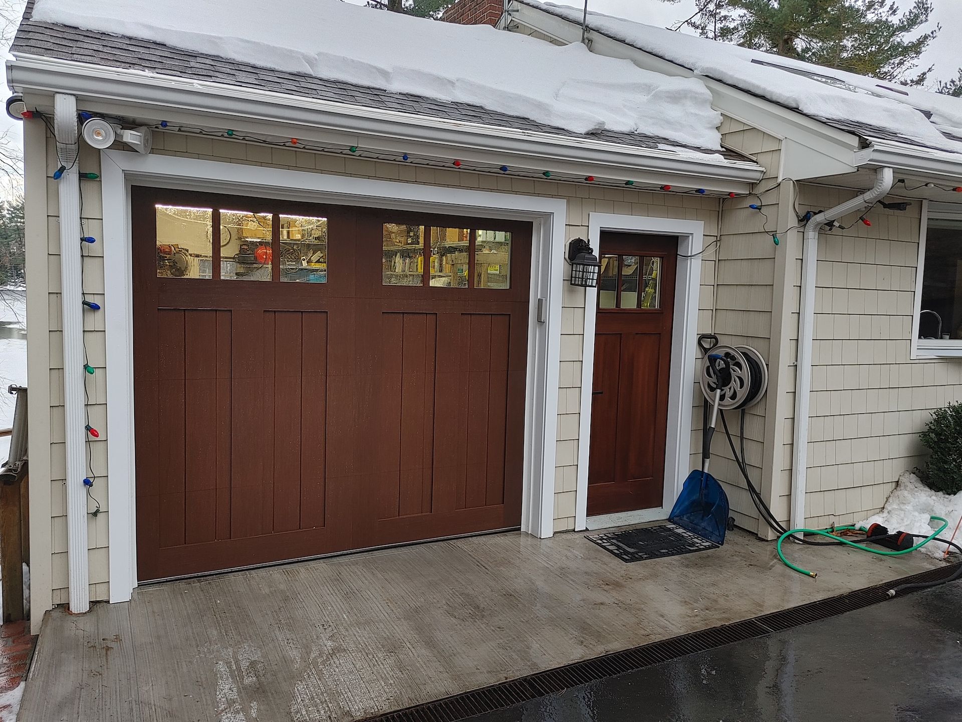 Snowy driveway with a brown garage door, side entry door, and wet concrete patio.