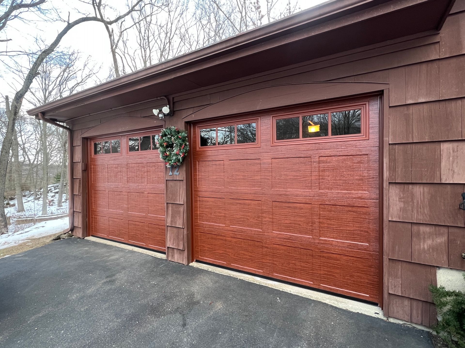Two reddish-brown garage doors on a brown house beside a driveway, with snow visible nearby.
