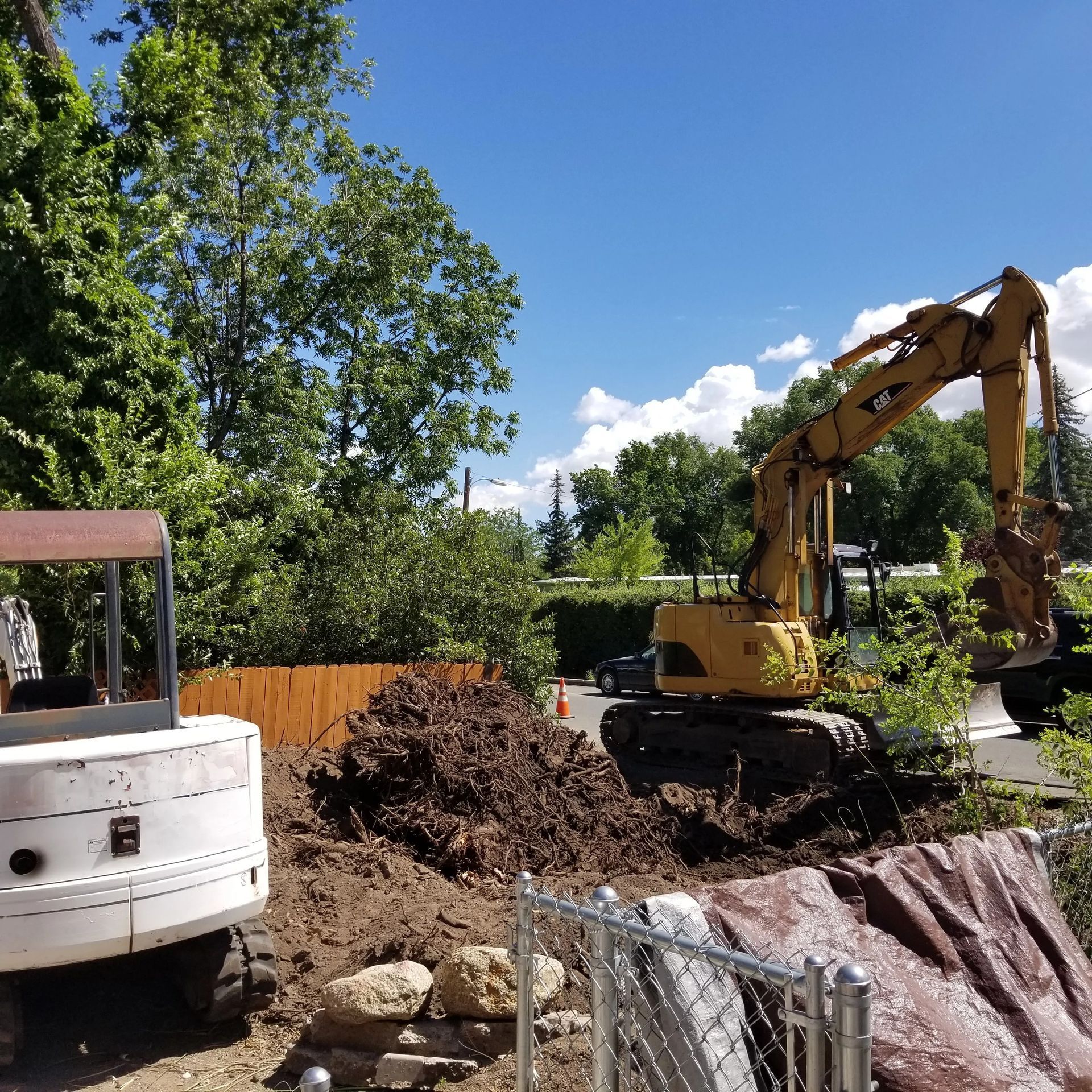 Construction site with excavator and small digger, digging in front of a fence, under a blue sky.