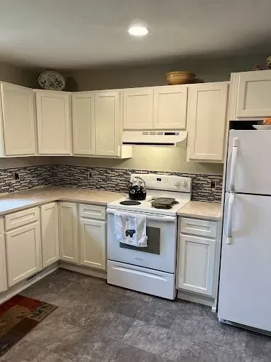 A kitchen with white cabinets, a white stove and refrigerator, a gray tile backsplash, and light countertops.