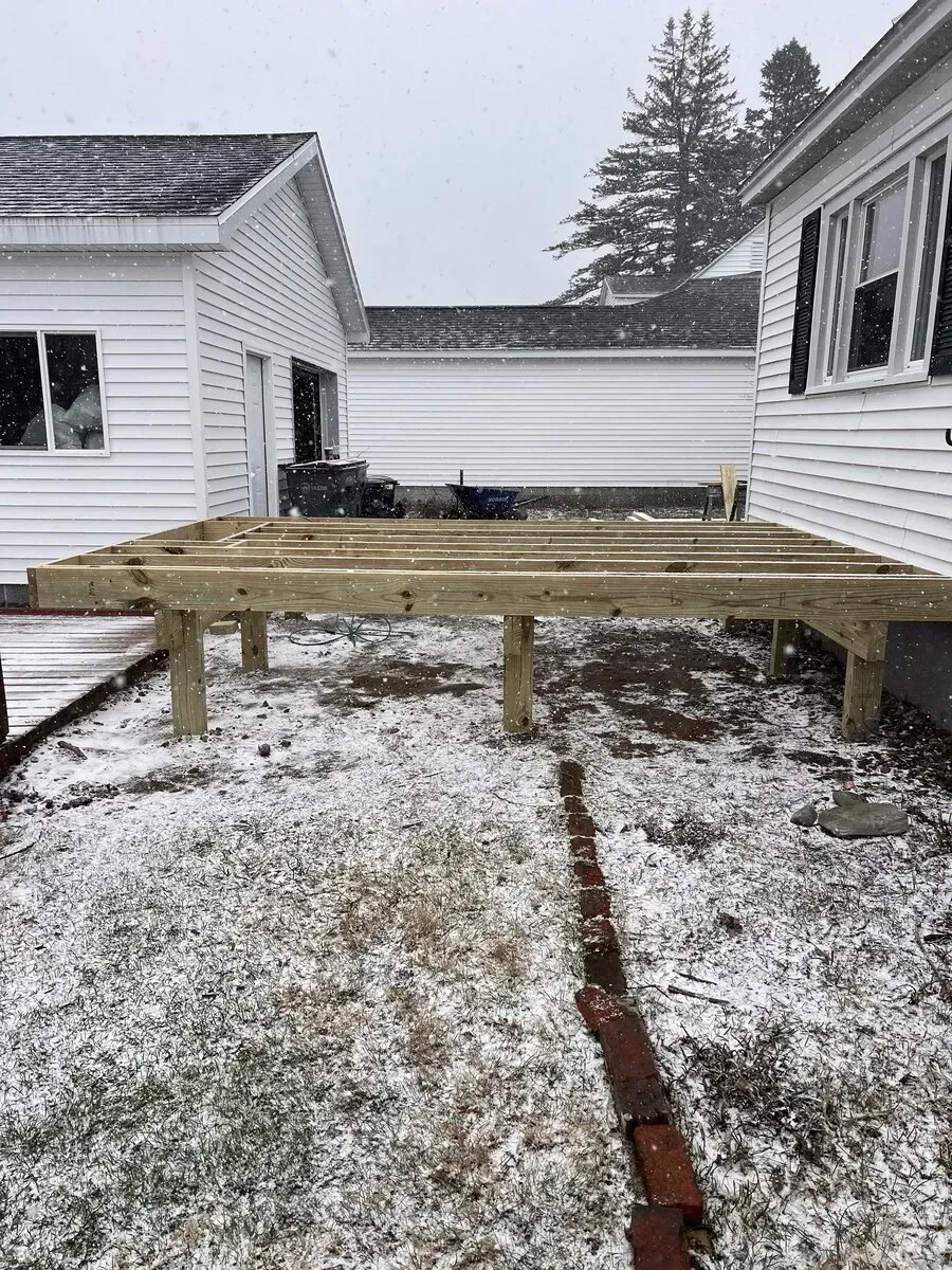 Unfinished wooden deck frame attached to a house exterior during a light snowfall.