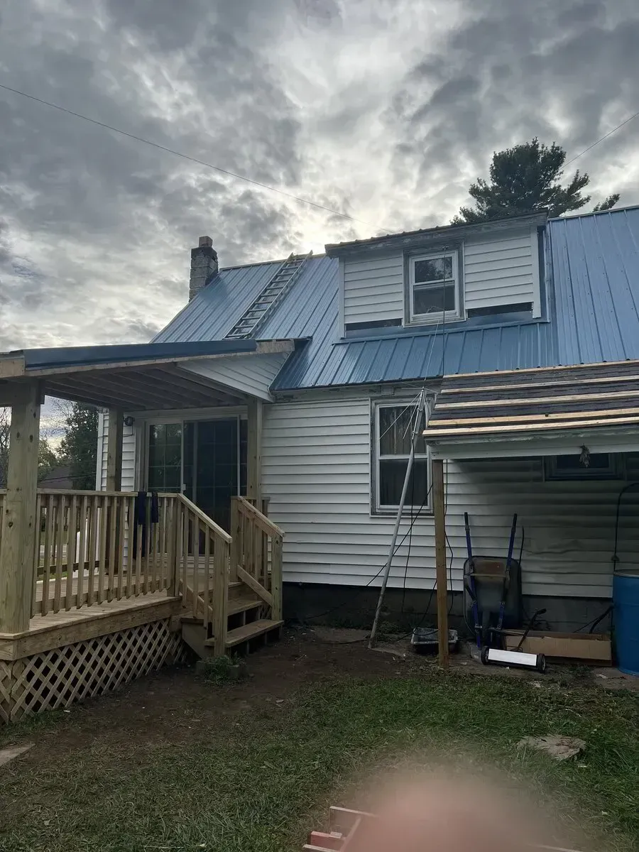 A white house with a blue metal roof, a wooden porch, and an unfinished addition against a cloudy sky.
