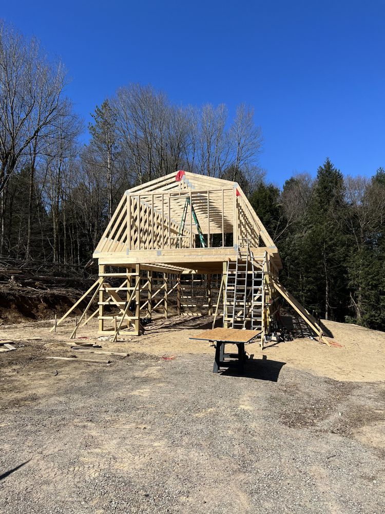 A partially constructed wooden barn frame stands on a gravel lot under a clear blue sky, surrounded by bare trees.
