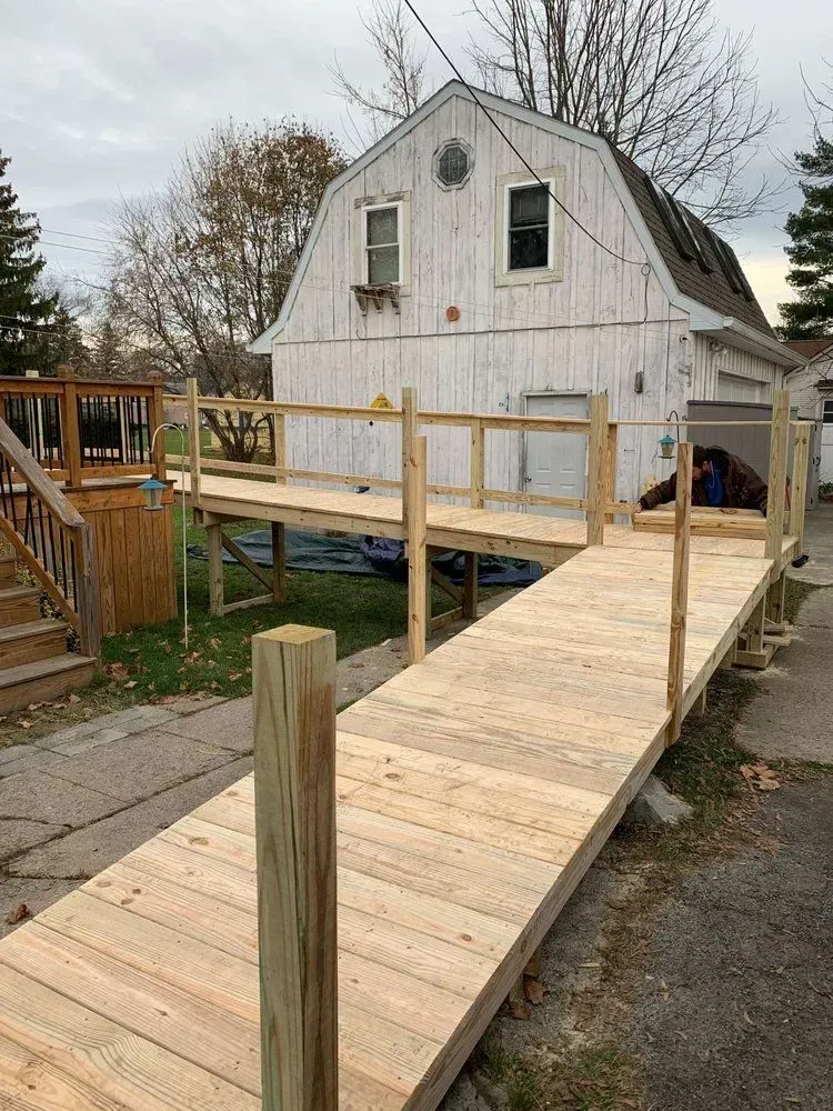 A wooden wheelchair ramp leading from a paved driveway to a residential deck, set in front of a white barn-style garage.