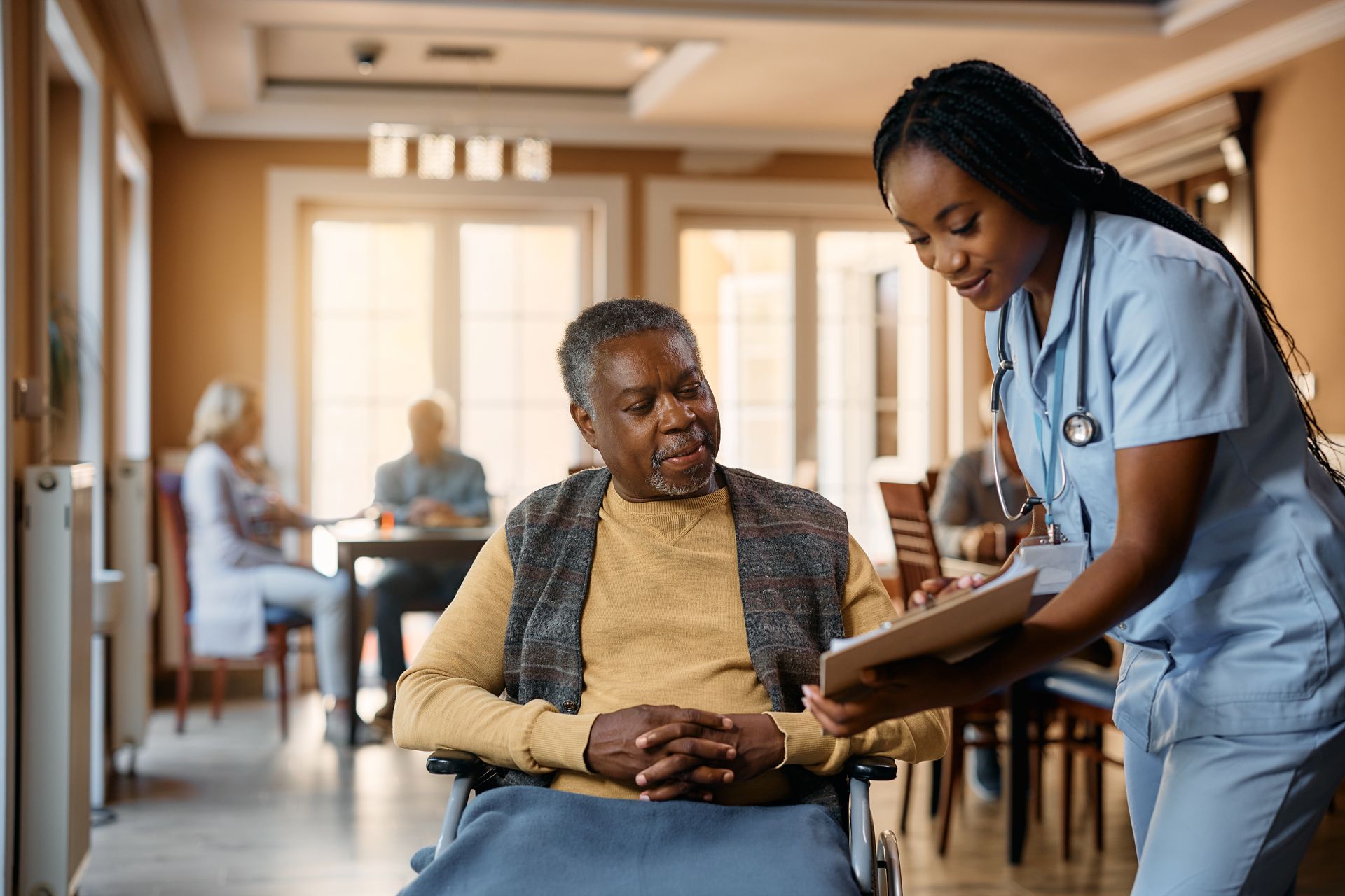 Nurse in blue scrubs reviews chart with Black man in wheelchair, sunlit interior.
