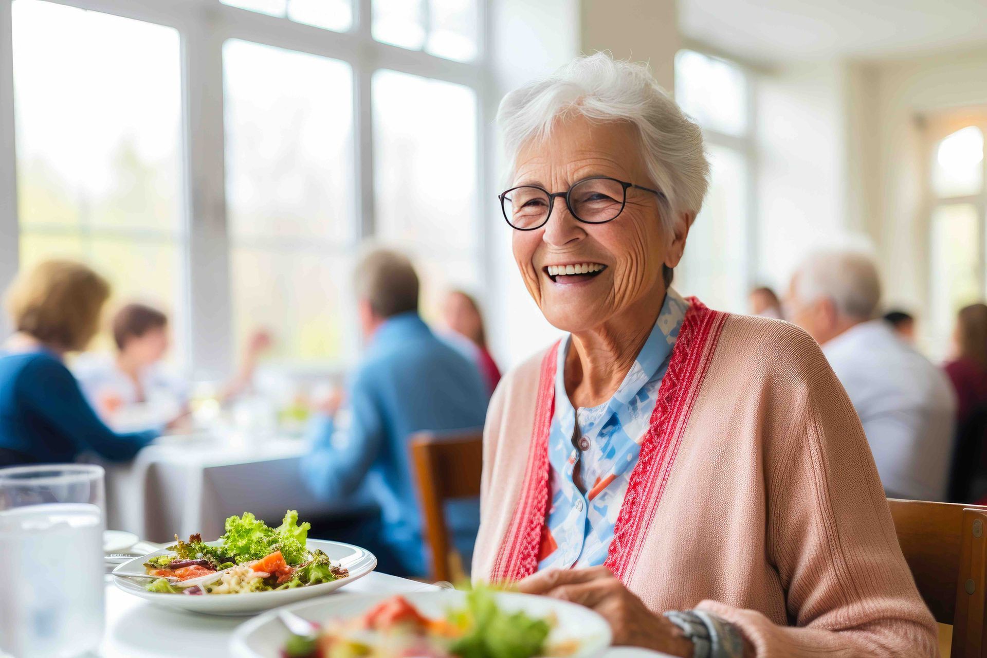 Smiling elderly woman wearing glasses, seated at a table in a dining room, eating salad.