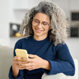 Woman with curly gray hair, wearing glasses, looks at phone, smiles. Contact Solution Web Designs For A Free Consultation. Your Partner in Building a Thriving Acupuncture Practice.