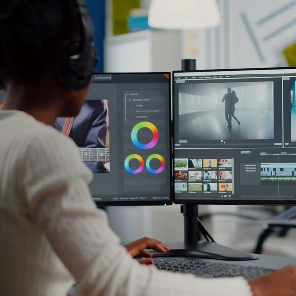 Person editing video on a dual monitor setup, color correction tools, and video timeline visible.