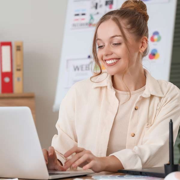 A smiling person in a cream-colored shirt works on a laptop at a desk with binders and a digital drawing tablet nearby.