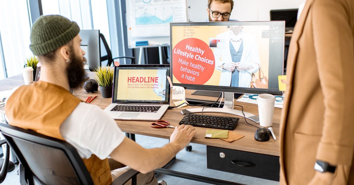 A web design Long Island person in a beanie sits at a desk with a laptop and monitor, collaborating with a colleague in an office.