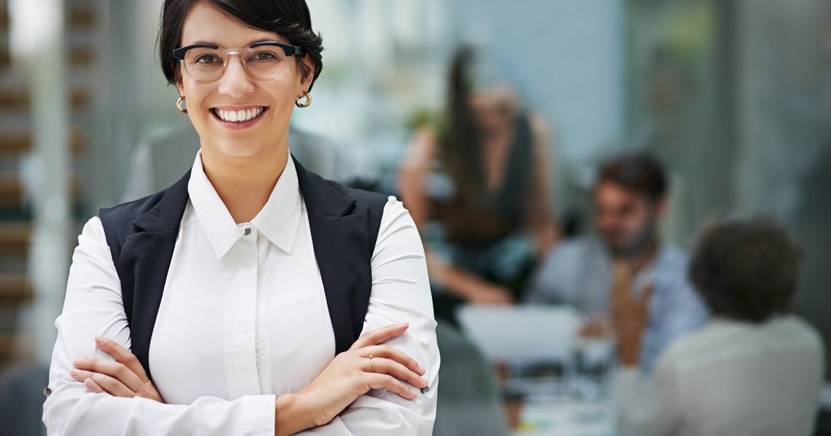 A woman in glasses smiles, arms crossed, standing in front of a blurred meeting in an office setting on Websites for Accountants and Accounting Firms by Solution Web Designs