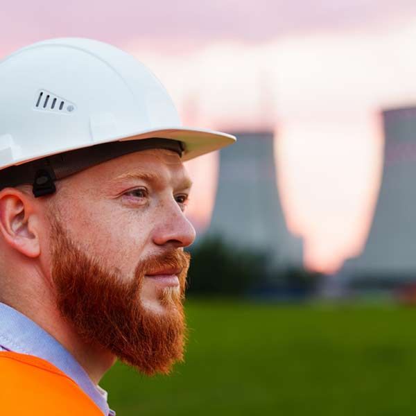Nuclear Engineer with red beard, wearing a white hard hat and orange vest, looking off-camera; industrial buildings in the background.