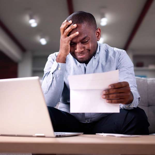 A man looking stressed while reading a Pay Per Click invoice near a laptop. Indoors, neutral colors.