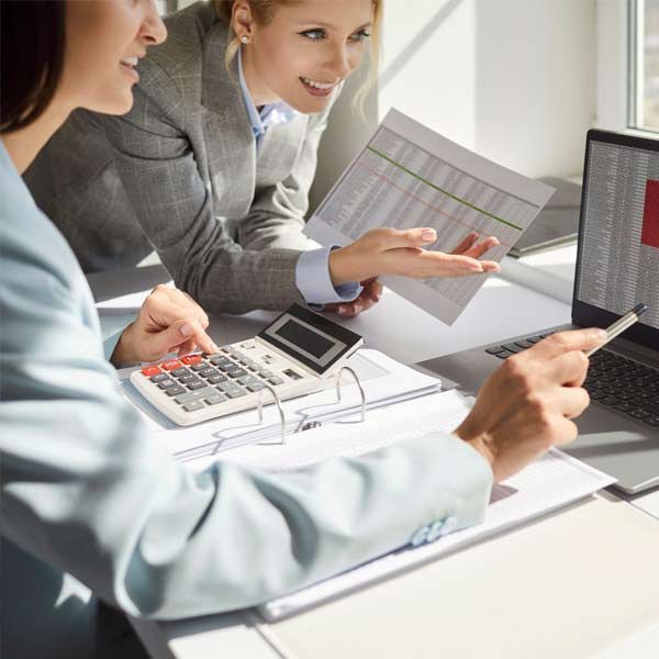 Two female accountants in business suits are reviewing data on a laptop and paper documents, using a calculator.