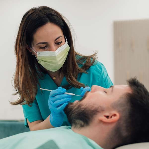 Dentist examining a patient's teeth. Dentist wears a mask, gloves, and scrubs. Patient reclines in a dental chair.