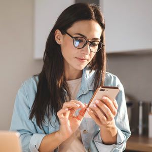 Woman doctor wearing glasses looks down at a smartphone, holding it in both hands, in a kitchen setting decided to Contact Solution Web Designs For A Free Consultation on a Website for Integrative and Functional Doctors
