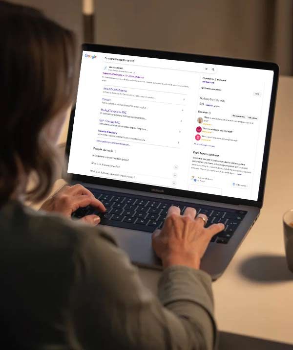 Person typing on a laptop, searching Google for an integrative and functional medicine doctor. Well-lit indoor setting.