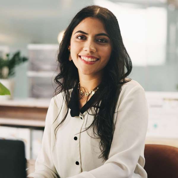 An insurance broker with dark hair, smiling, wearing a white shirt with a black collar, in an office setting.