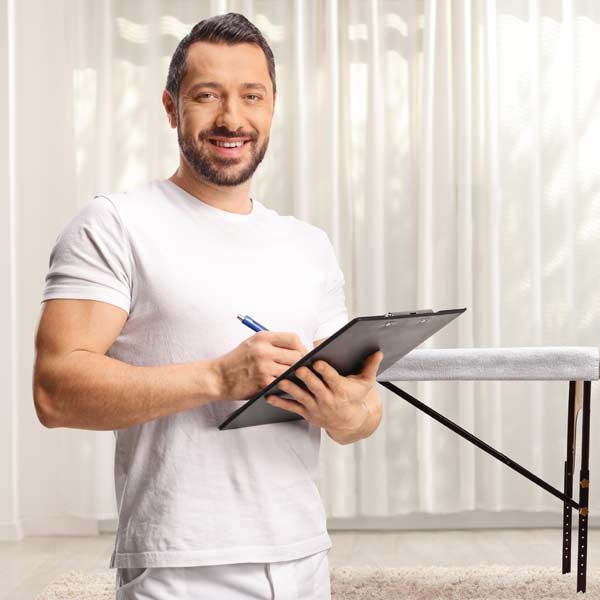 Massage Therapist in white shirt smiling, writing on a clipboard, massage table in the background.
