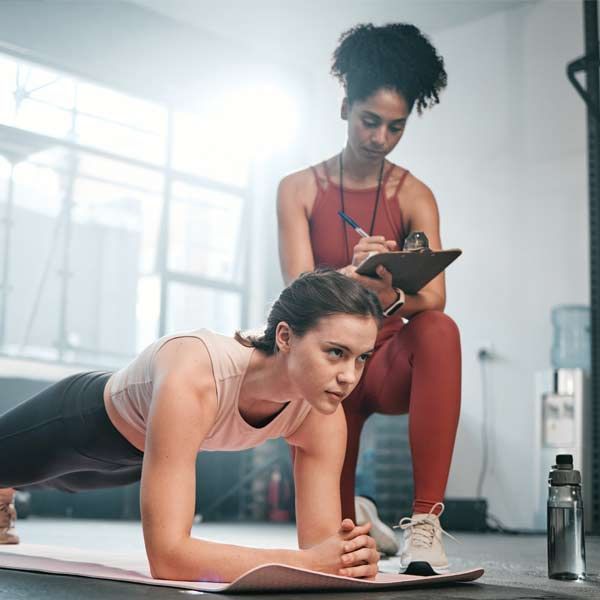 A woman in plank pose, assisted by a personal trainer, writing on a clipboard in a gym setting.