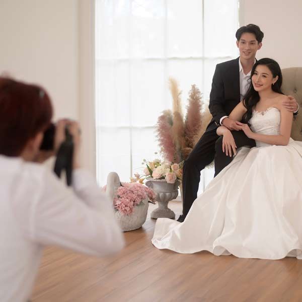 A photographer takes a photo of a couple in wedding attire; the woman in a white dress, the man in a black suit, indoors.