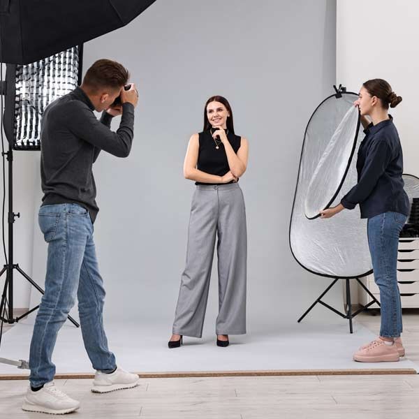 A photographer is taking a photo of a professional model with an assistant holding a reflector in a studio with lighting equipment.